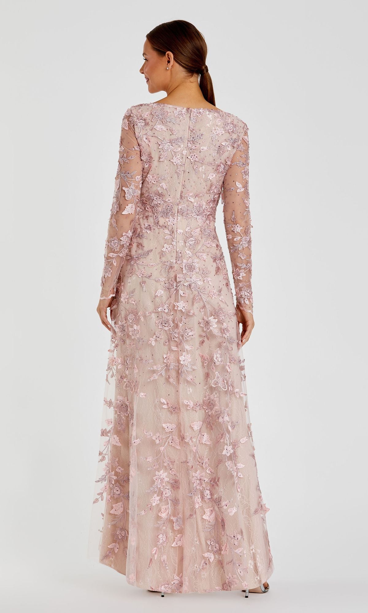 A woman faces away, wearing the Mac Duggal 70224 Long Sleeve Long Formal Dress in light pink with floral embroidery and a sheer, floor-length overlay.